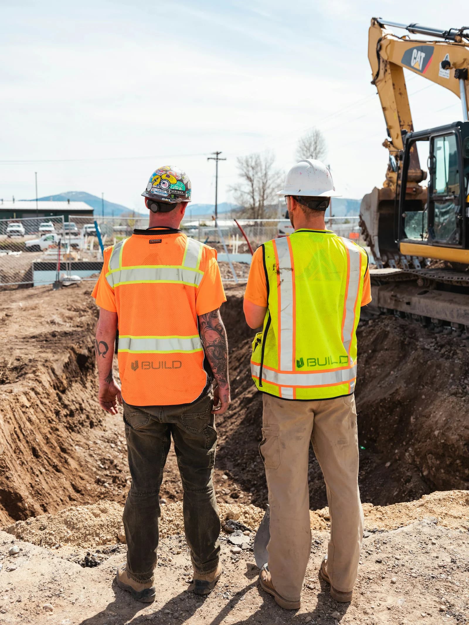 Two U Build crew members in safety vests overlooking an excavation beside an excavator